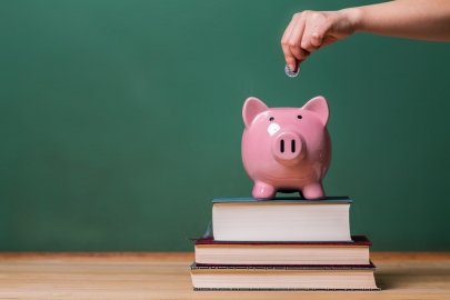Piggy bank sitting on top of a small stack of books with a hand above it about to drop a coin in.
